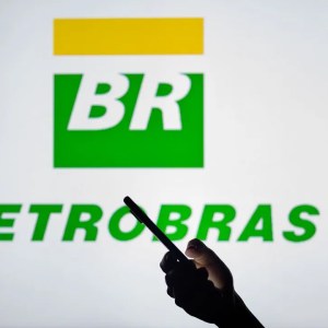 A foto mostra o logo da empresa Petrobras no plano de fundo, com uma pessoa no primeiro plano segurando um celular.