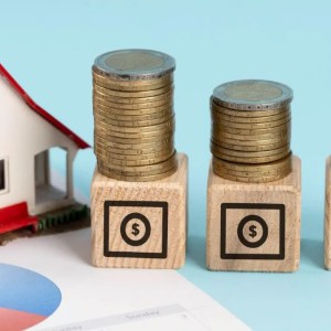 Foto de blocos de madeira com moedas empilhadas e uma casa ao lado. A matéria destaca quanto renderam R$ 1 mil nos maiores fundos imobiliários na bolsa de valores