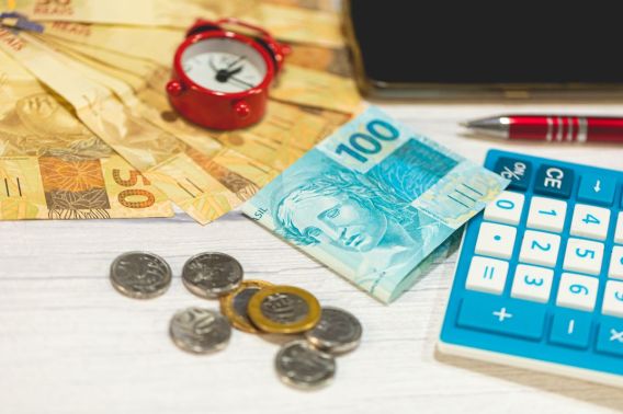 Imagem para a matéria sobre quanto rendem 4 milhões na poupança em que aparecem notas de 50 reais, 100 reais e moedas em cima de uma mesa. Na mesa também estão uma calculadora, um celular e uma caneta.