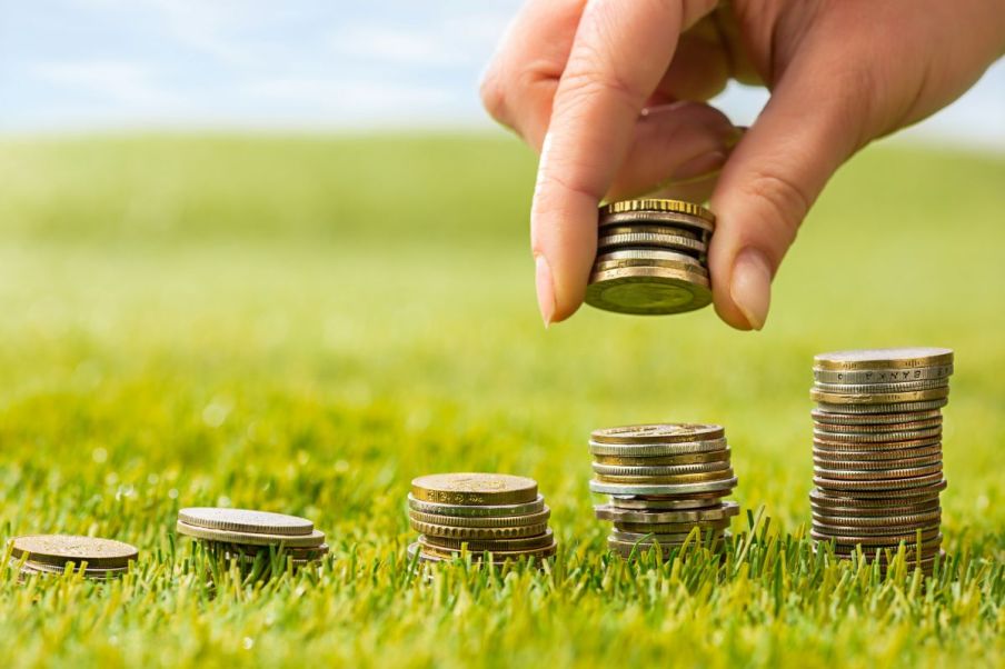 Imagem para matéria sobre fundos do agronegócio em que aparecem várias pilhas de moedas e uma mão empilhando mais moedas. Essas moedas estão em um campo verde.