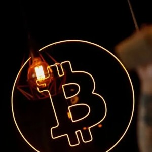 Foto do símbolo do bitcoin, com uma moeda laranja e uma letra B dentro. Ao fundo, há o perfil desfocado de um homem tomando um café.