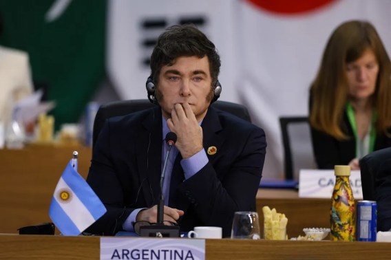 Foto de um homem com cabelo escuro, vestindo um terno. Em frente, uma bandeira pequena da Argentina.