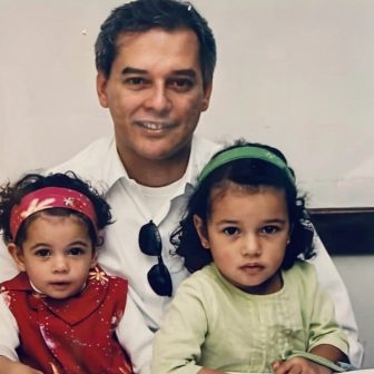 Foto de um homem vestindo uma camisa branca. Ele está com duas meninas, pequenas, junto dele.