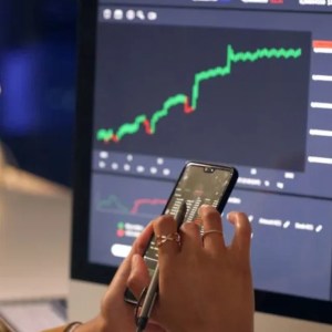Imagem de uma mulher olhando para o celular, verificando uma lista. Em frente a ela, um computador com gráficos e informações do mercado financeiro.