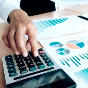 Imagem em que aparece uma mão de uma pessoa branca mexendo em uma calculadora que está em cima de uma mesa de madeira. Ao lado da calculadora, papéis com gráficos.