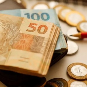 Imagem de um bolo de notas de 50 reais por cima de um bolo de notas de 100 reais. Ambos estão por cima de uma carteira preta. Ao redor, várias moedas de 1 real e 50 centavos. Junto a esses objetos tem um lápis vermelho e um caderno.