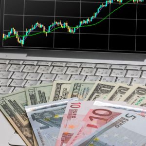 Imagem de um computador com gráfico na tela. Em cima do teclado, notas de dólar e euro.