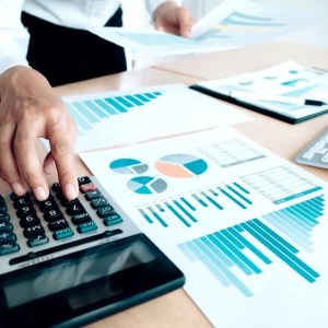 Imagem em que aparece uma mão de uma pessoa branca mexendo em uma calculadora que está em cima de uma mesa de madeira. Ao lado da calculadora, papéis com gráficos.