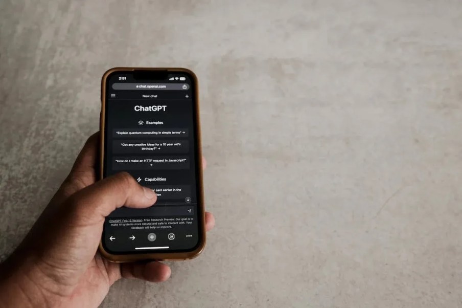 Imagem da mão de uma pessoa branca segurando um celular. Na tela está a plataforma de inteligência artificial ChatGPT.