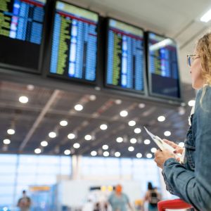 Imagem de uma mulher branca e loira de óculos olhando para o painel de decolagens de um aeroporto.