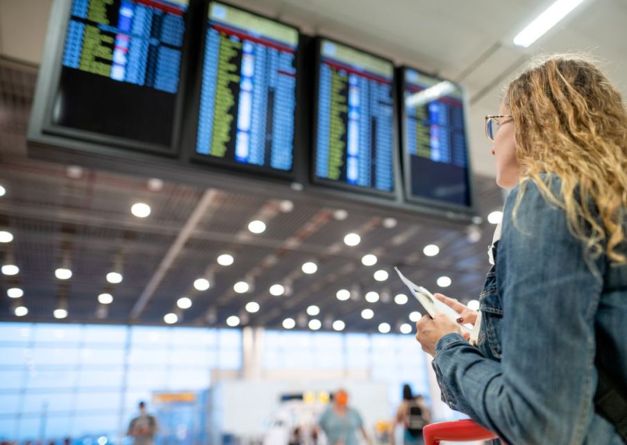 Imagem de uma mulher branca e loira de óculos olhando para o painel de decolagens de um aeroporto.