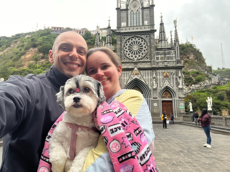 Foto de um casal formado por um homem e uma mulher. Ele estão segurando um cachorro.